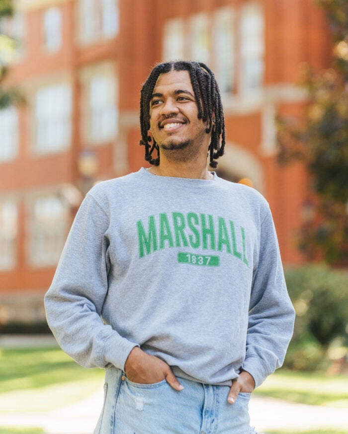 Kameron smiles and looks to the side while wearing a Marshall crewneck while standing in front of Marshall University's Old Main.