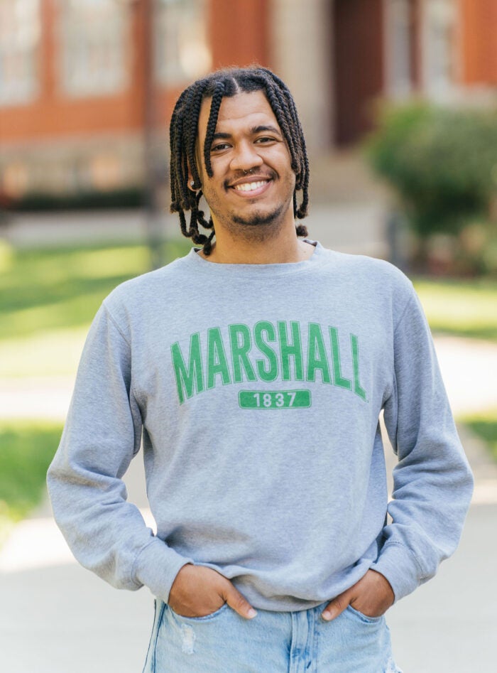 Cameron Riggins looks at the camera and smiles while wearing a Marshall shirt and standing in front of Marshall University's Old Main