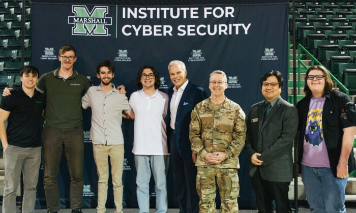 Brandon Anderson and a group of students stand with Brad D. Smith and additional CyberCon speakers in front of a Marshall University Institute for Cyber Security banner