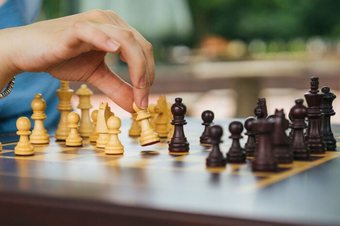 Close-up of a student’s hand moving a light-colored chess piece on a chessboard during an outdoor game.