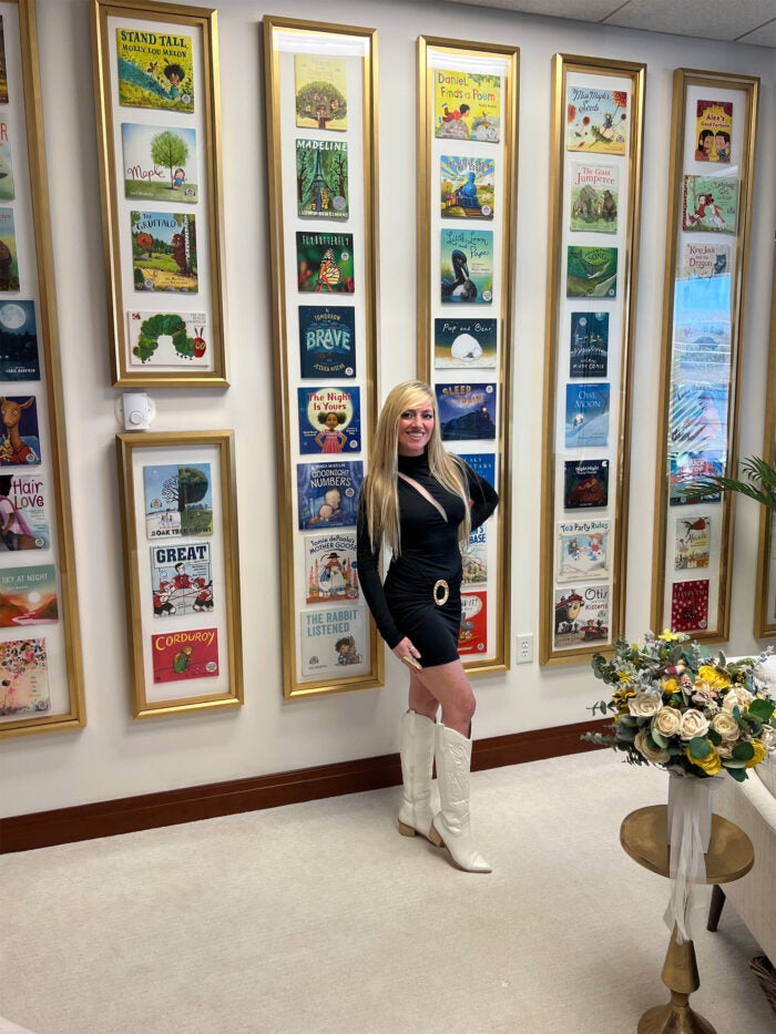 Jaimie Lima wears a black dress and white boots while standing in front of a wall of framed book covers from Dolly's Imagination Library