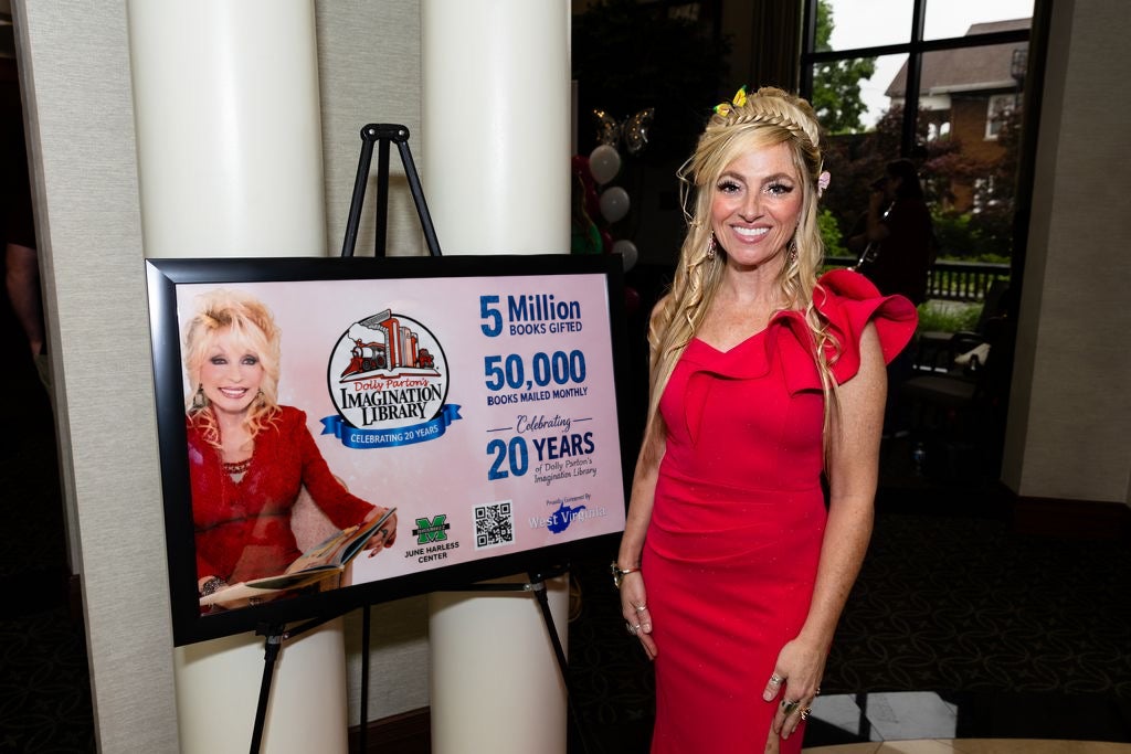 Jamie Lima wears a red dress and stands beside a poster celebrating 20 years of Dolly Parton’s Imagination Library in West Virginia during the Harless Hall of Fame dinner and awards ceremony at Marshall University's Brad D. Smith Foundation Hall