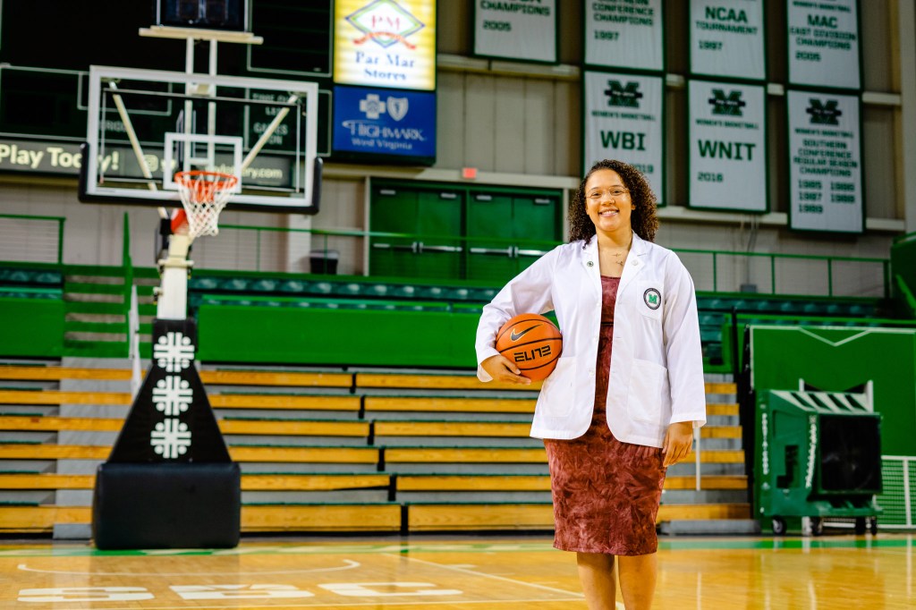 Taylor Pearson, former Herd Women's Basketball player and current third-year medical student at Marshall University's Joan C. Edwards School of Medicine, stands on the basketball court wearing her white coat and holding a basketball