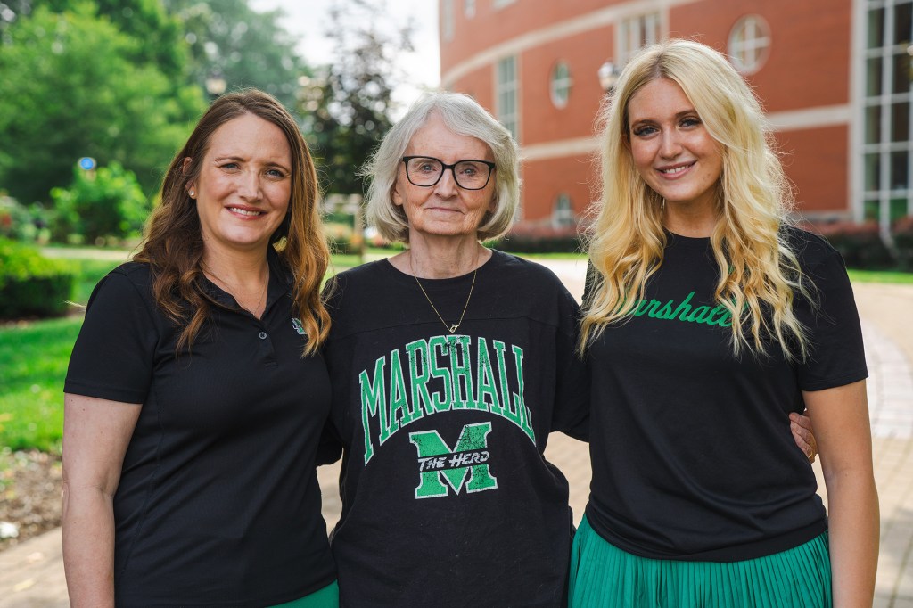 Three generations of Marshall University women stand together on campus, wearing black and green Marshall attire.