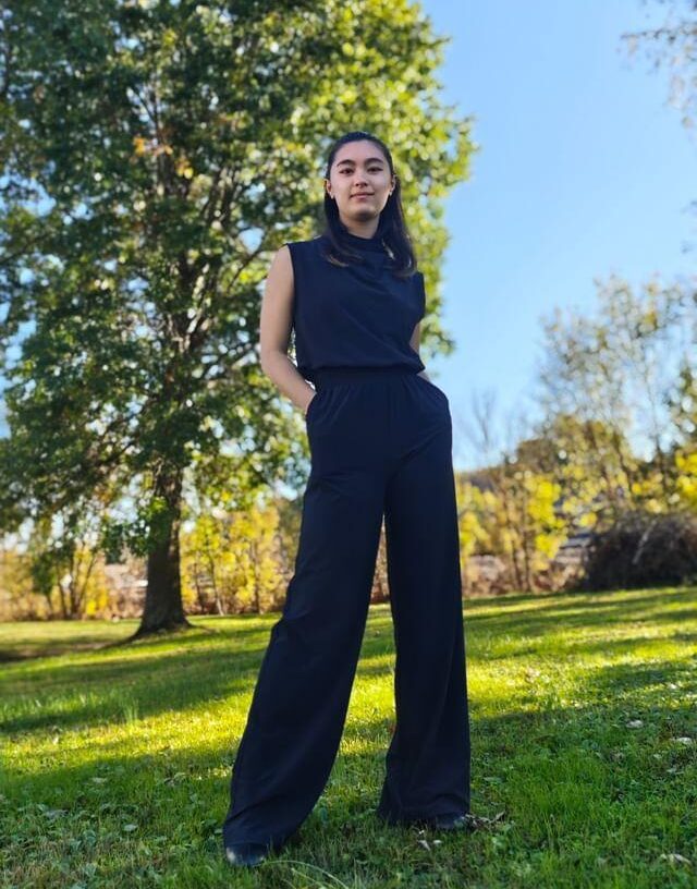 Sarah Talkington wears a black jumpsuit and smiles at the camera with a large tree behind her