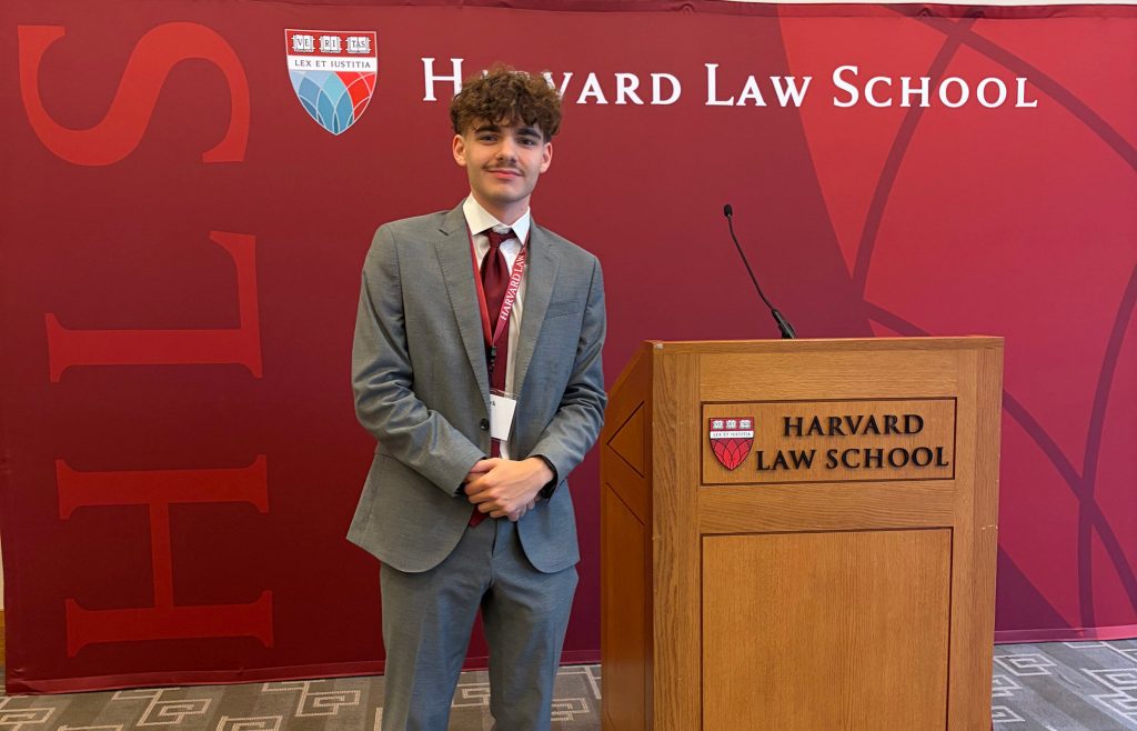 Mark Hysell wears a suit and smiles next to a Harvard Law School podium in front of a Harvard backdrop