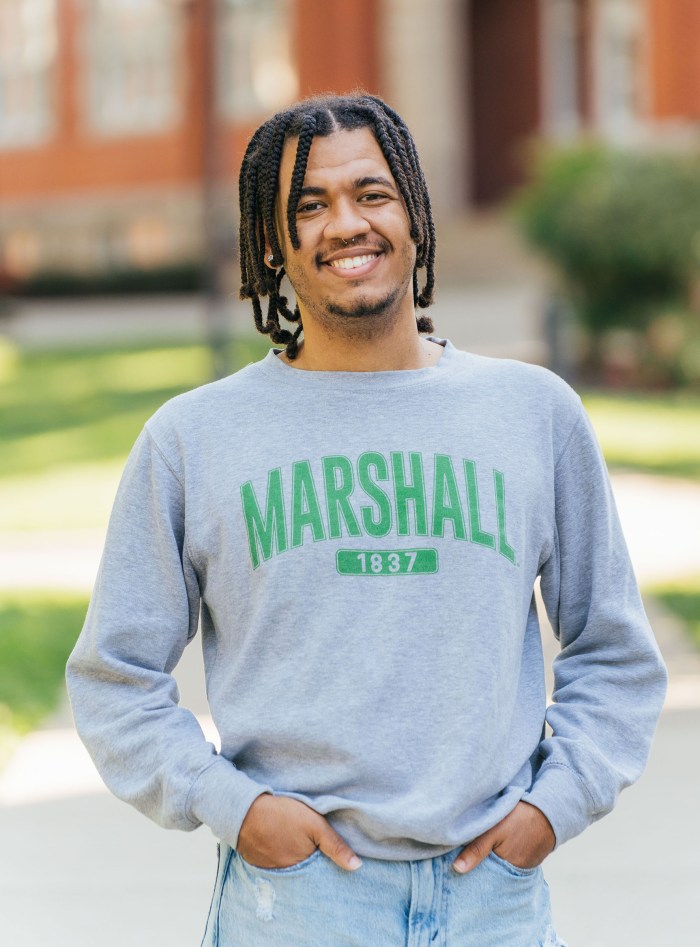 Cameron Riggins looks at the camera and smiles while wearing a Marshall shirt and standing in front of Marshall University's Old Main