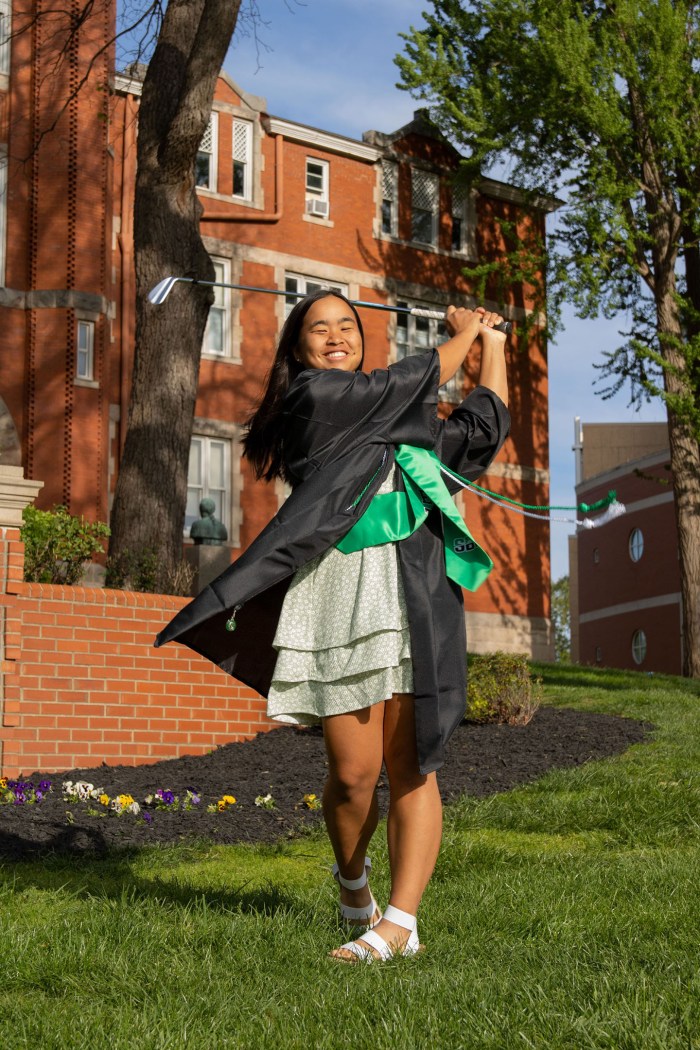 Hanna Shrout wears her graduation gown and swings a golf club in front of Marshall University's Old Main