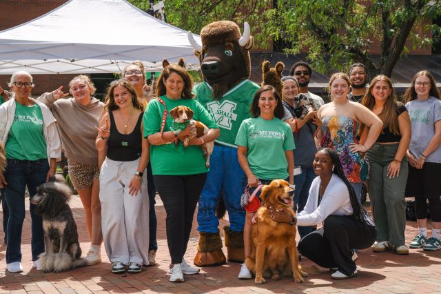 A large, diverse group of people pose outside on the MSC Plaza with Marco, the Marshall University's bison mascot. Several people hold or sit with dogs of different sizes, with a white canopy tent and trees in the background.