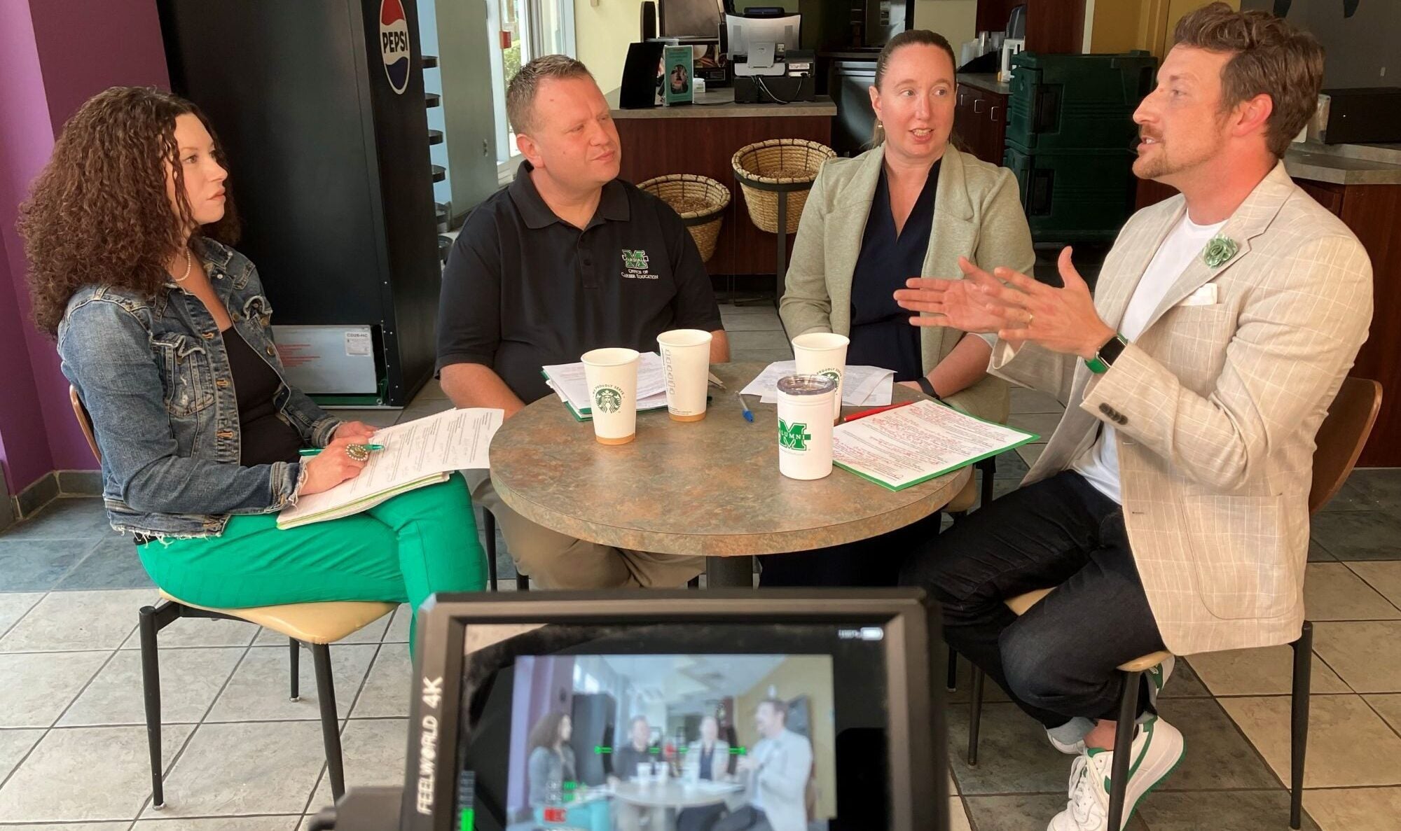 4 people sitting around a table having a discussion with a camera screen in front recording the moment.