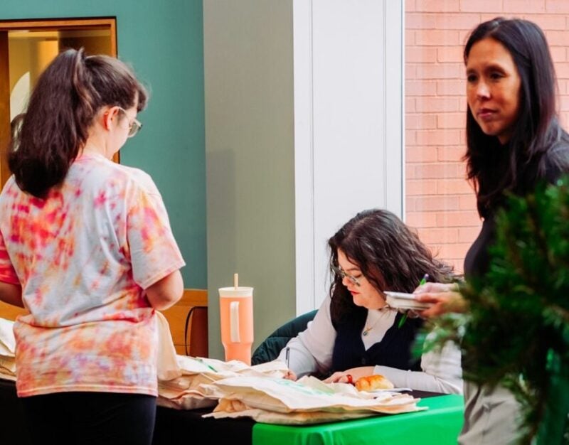 A seated team member assists a student at a Marshall Online welcome table stocked with tote bags and giveaways, as another staff member stands nearby.