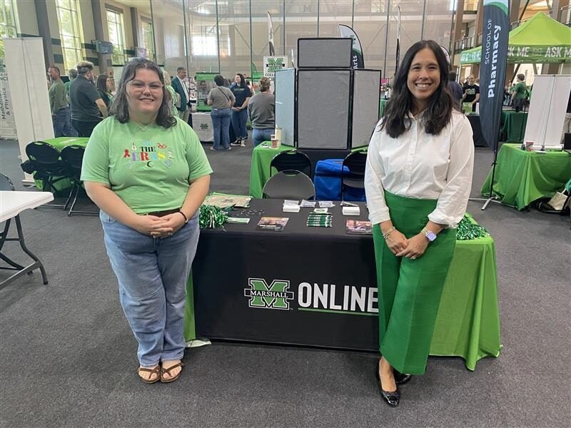 Two Marshall Online team members smile beside a Marshall Online table at a campus event, with green giveaways and flyers on display and other booths in the background.