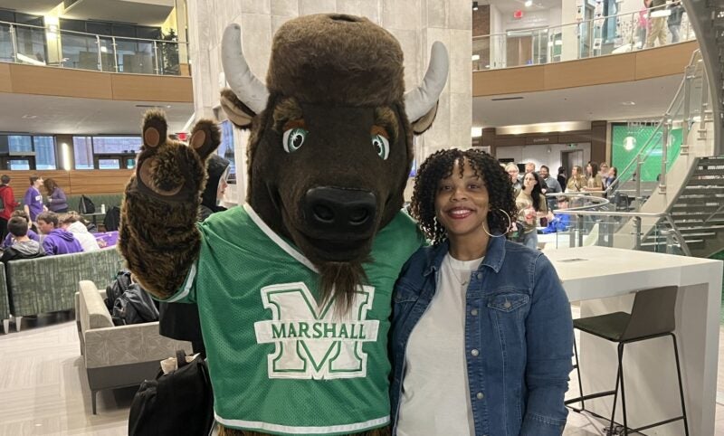 Marco, Marshall University's buffalo mascot in a green Marshall jersey, waves while posing with a smiling woman in a campus building.