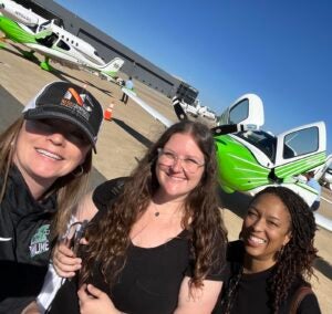 Three people smile for a selfie on an airport tarmac with small propeller aircraft and a hangar in the background.