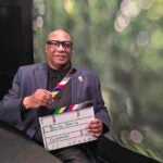 A presenter sits in a video studio holding a film slate labeled 
