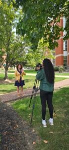 A videographer films a presenter in a yellow cardigan on a brick campus walkway, with trees and a campus building in the background.