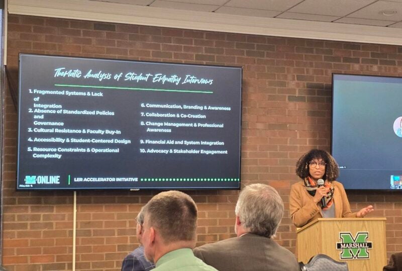 A presenter speaks at a Marshall-branded podium beside a screen titled 