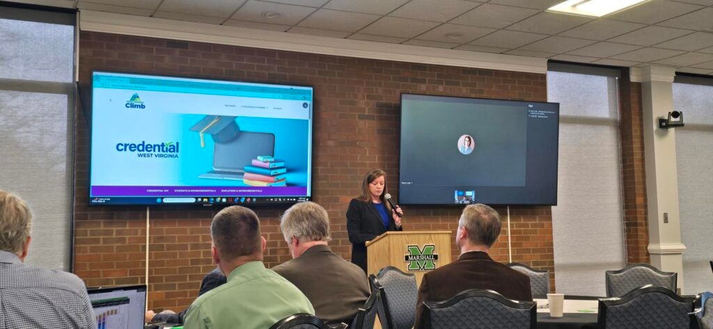 A presenter speaks at a Marshall-branded podium beside a screen displaying the Credential West Virginia website, as seated attendees look on.