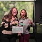 Two smiling women hold a film slate reading 