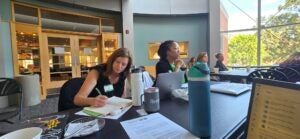 Attendees take notes and follow a presentation at a working session, seated at tables with laptops, notebooks and coffee.