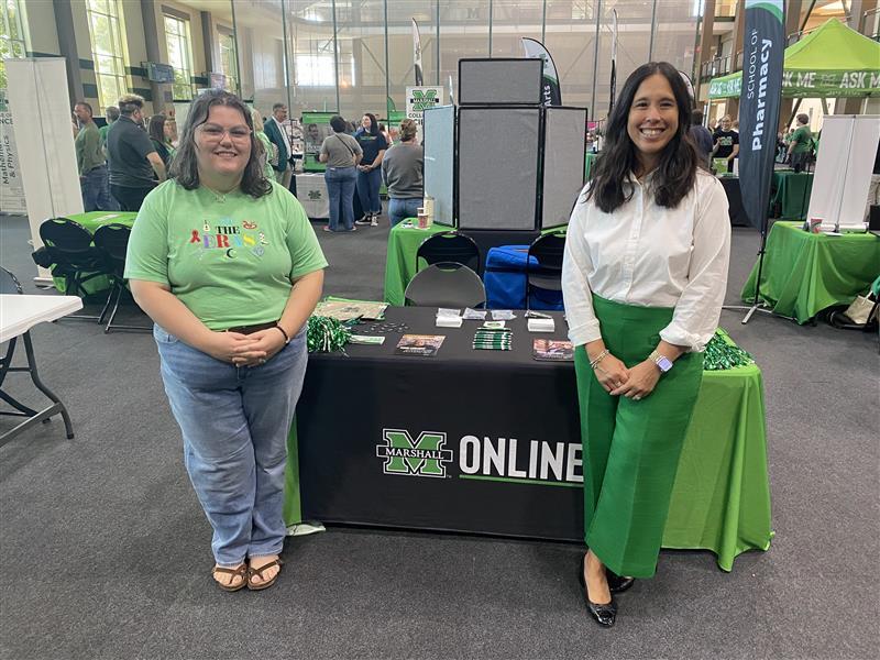 Two Marshall Online team members smile beside a Marshall Online table at a campus event, with green giveaways and flyers on display and other booths in the background.