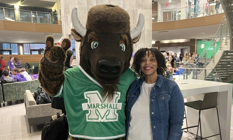 Marco, Marshall University's buffalo mascot in a green Marshall jersey, waves while posing with a smiling woman in a campus building.