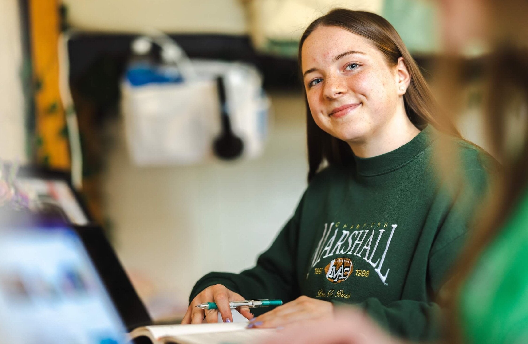 student studying at a laptop with an open textbook, smiling at the camera