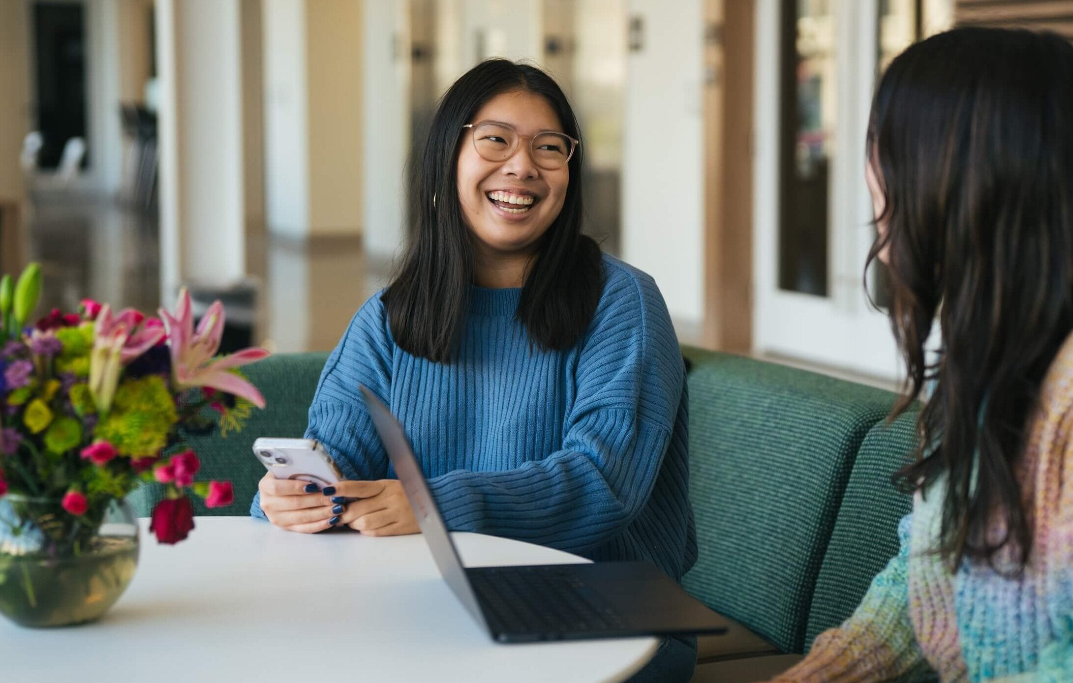 two students smiling at a table, one on a phone and one on a laptop