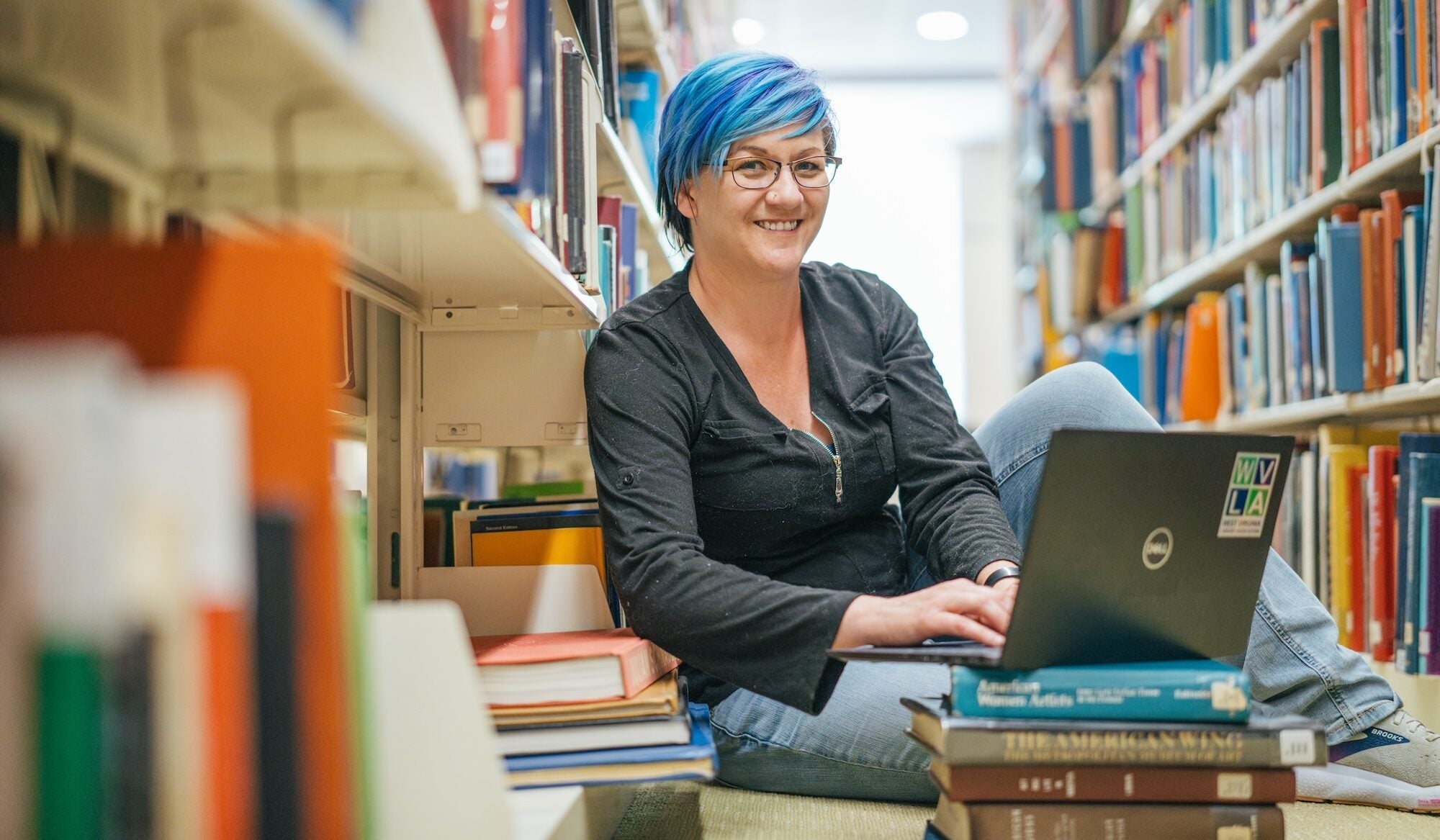 student sitting in between shelves in the library and working on a laptop