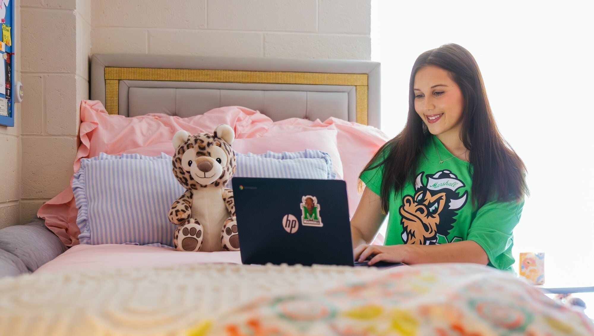 student working on a laptop in a residence hall on a bed, wearing a Marshall tshirt