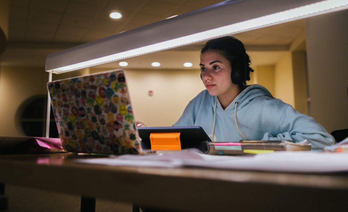 A student works on a laptop computer by the light of a desk lamp in a library