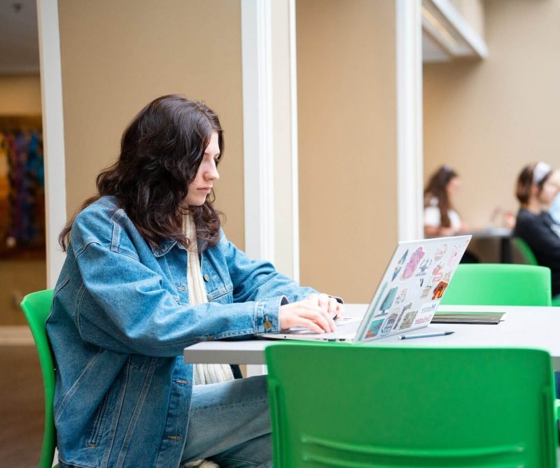 Student works on laptop in a building with other people around