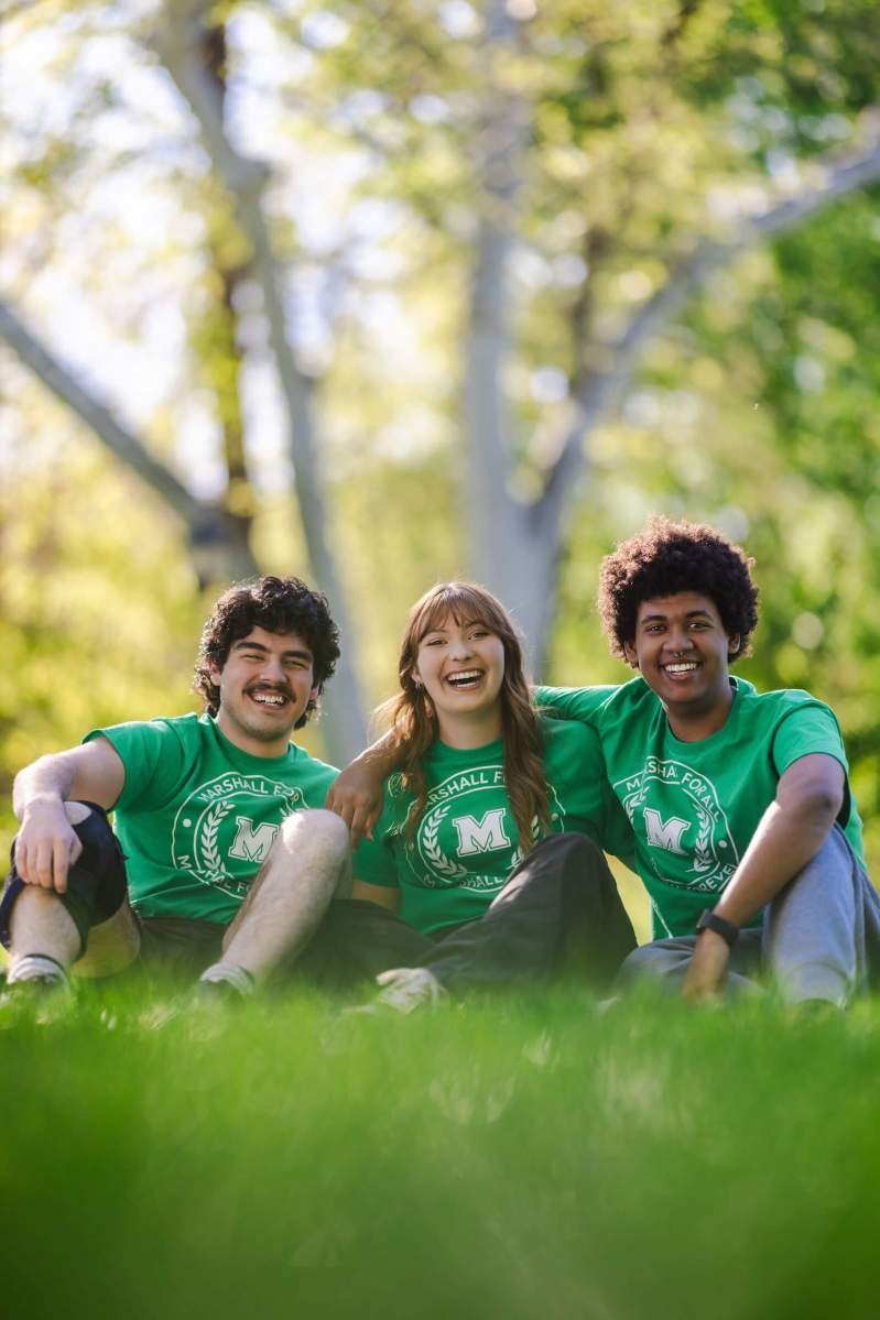 three students smiling at the camera and sitting on the grass outside the library