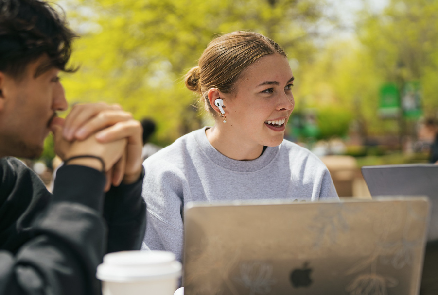 student with earbuds and laptop talks to other students