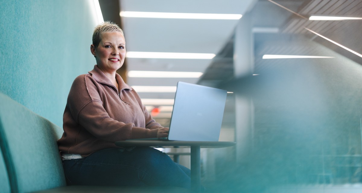 A student smiles at the camera while working on a laptop in a library common area