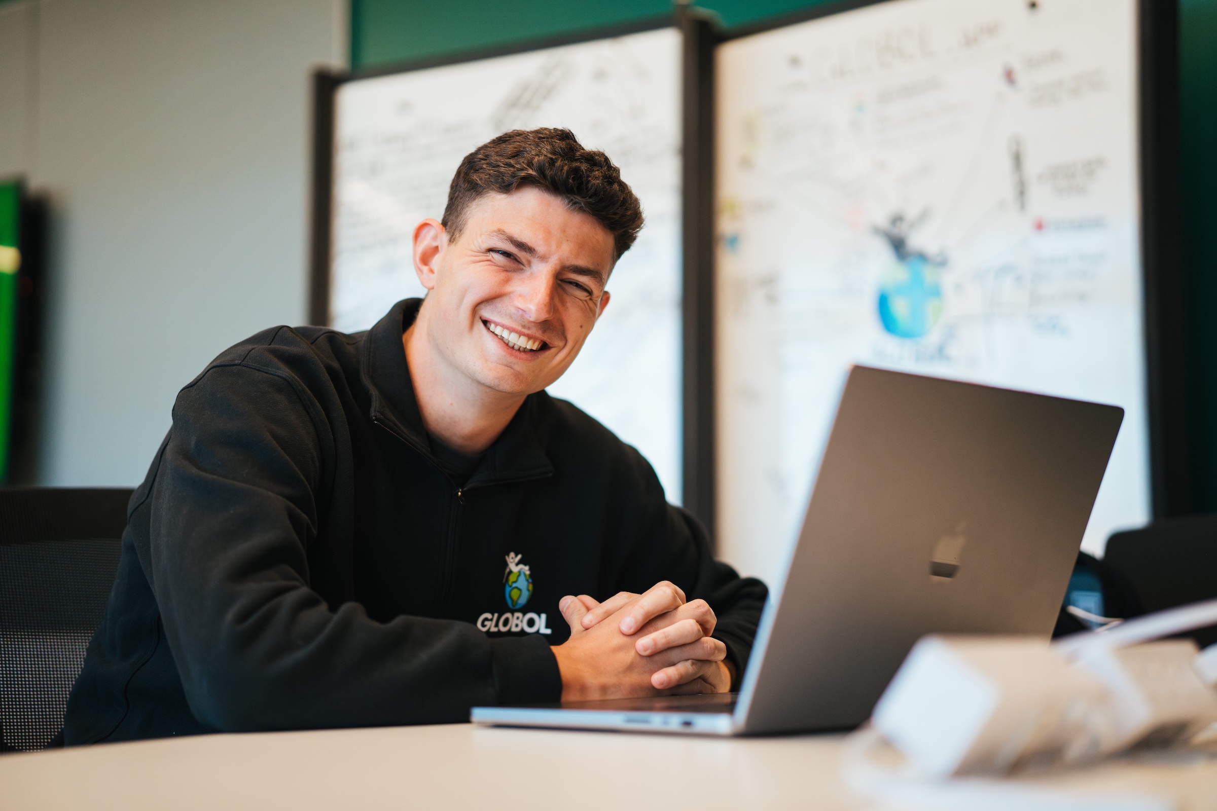 student smiling at the camera with a laptop in front of him and a white board in the background