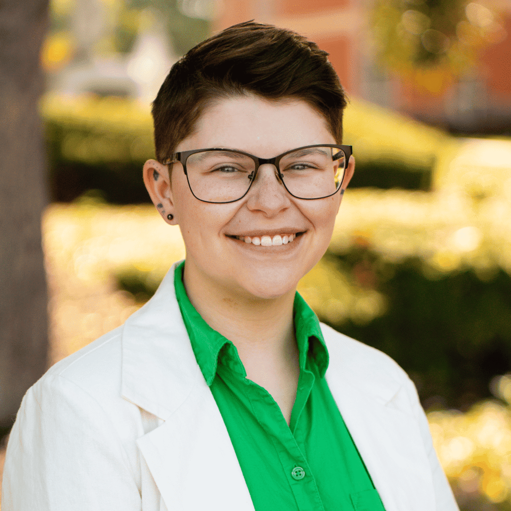 Headshot of a person with short brown haird and glasses wearing a green button up with a white blazer.