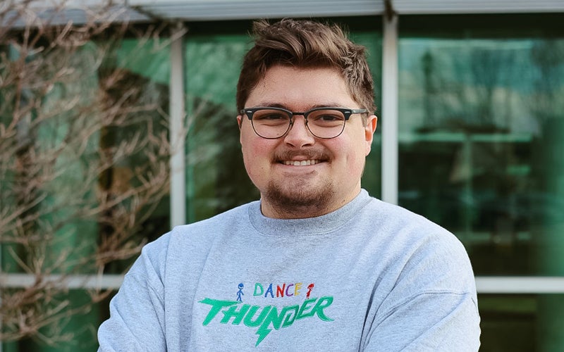Braden Cotton wears a Dance Thunder shirt while standing in front of Marshall's Harless Dining Hall