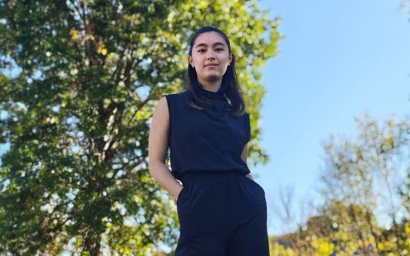 Sarah Talkington looks down at the camera while wearing a black jumpsuit and standing in a park