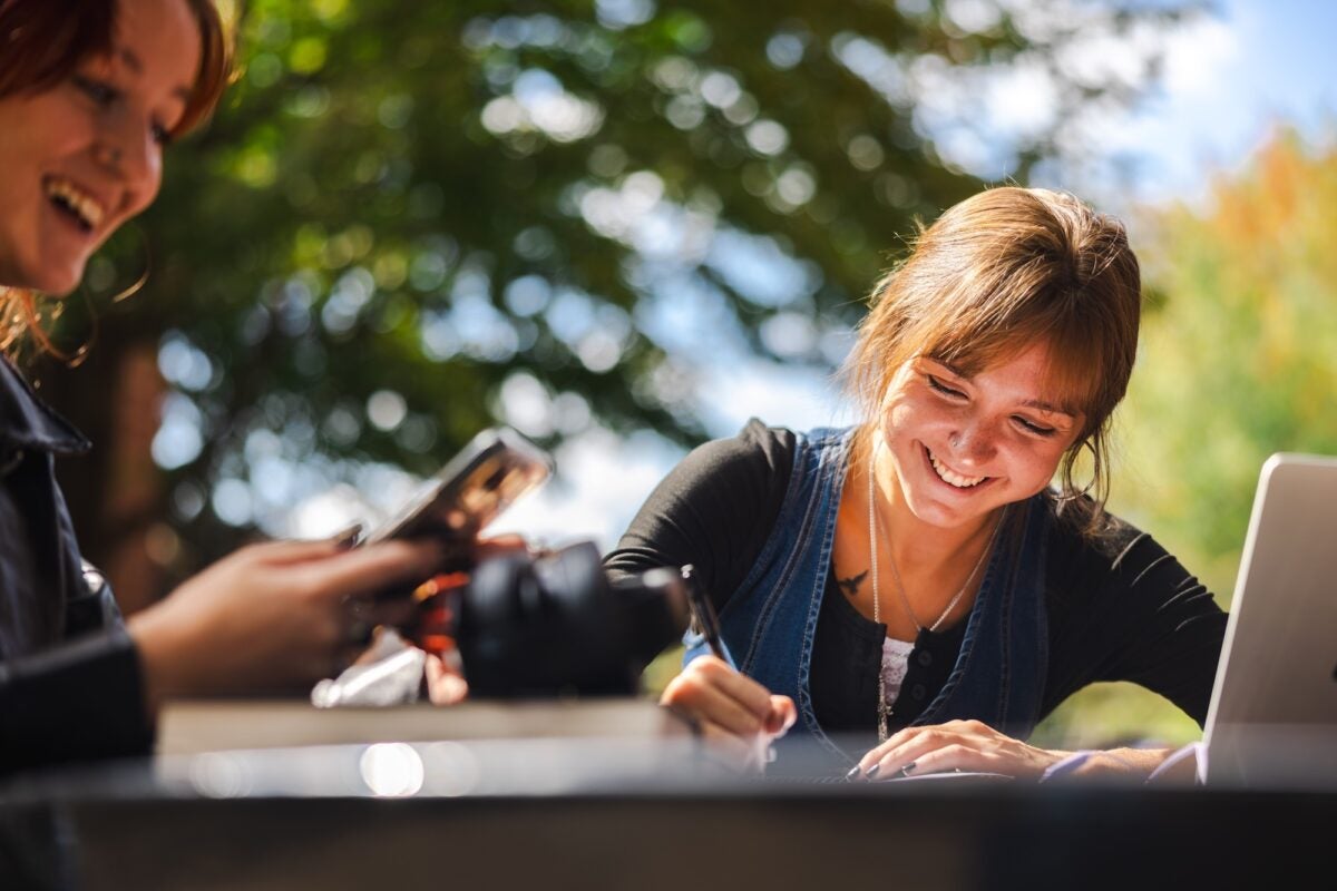 students studying outside at a table taking notes and using a phone and laptop