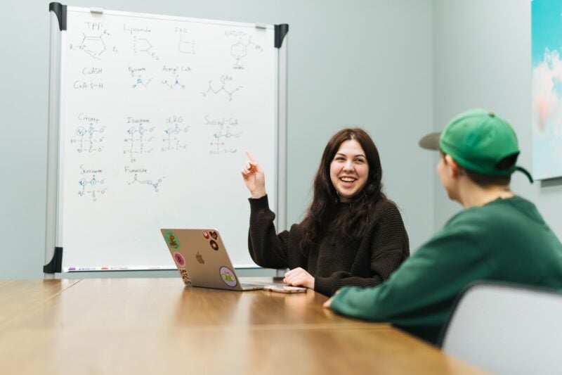 student on a laptop talking to another student and pointing at a white board