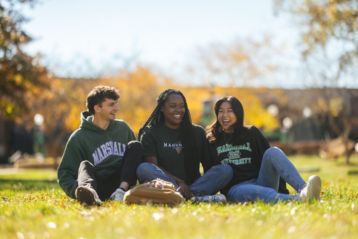 three students sitting in the grass on campus and laughing