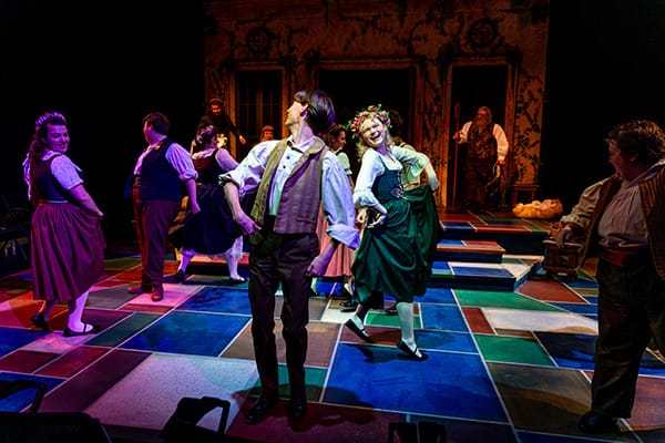 A troupe of actors in period costumes performs a lively scene on a stage with a colorful, stained-glass-like floor pattern.