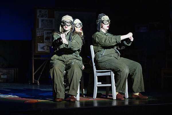 Three women in olive green flight suits and aviator goggles sit on chairs on a dimly lit stage, portraying pilots.