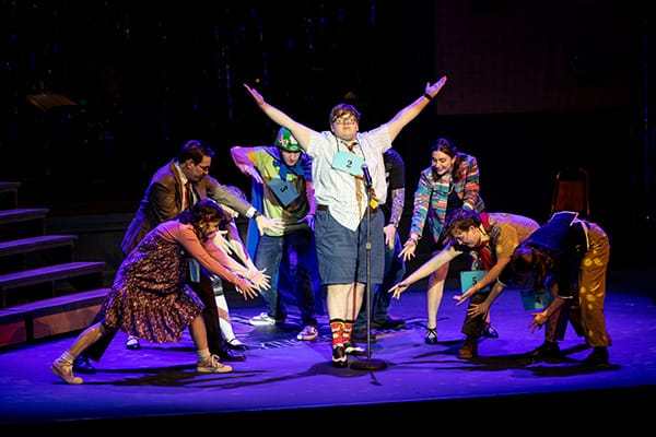 A group of actors on a dimly lit stage surround a central male performer with his arms raised in victory during a theatrical performance.