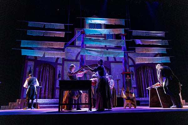 Actors in period costumes perform on a stage set resembling a print shop with hanging book pages under dramatic purple and blue lighting.