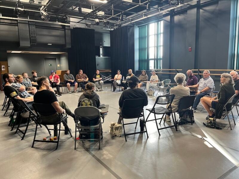 A group of people seated in a large circle during a discussion in a theater rehearsal space.