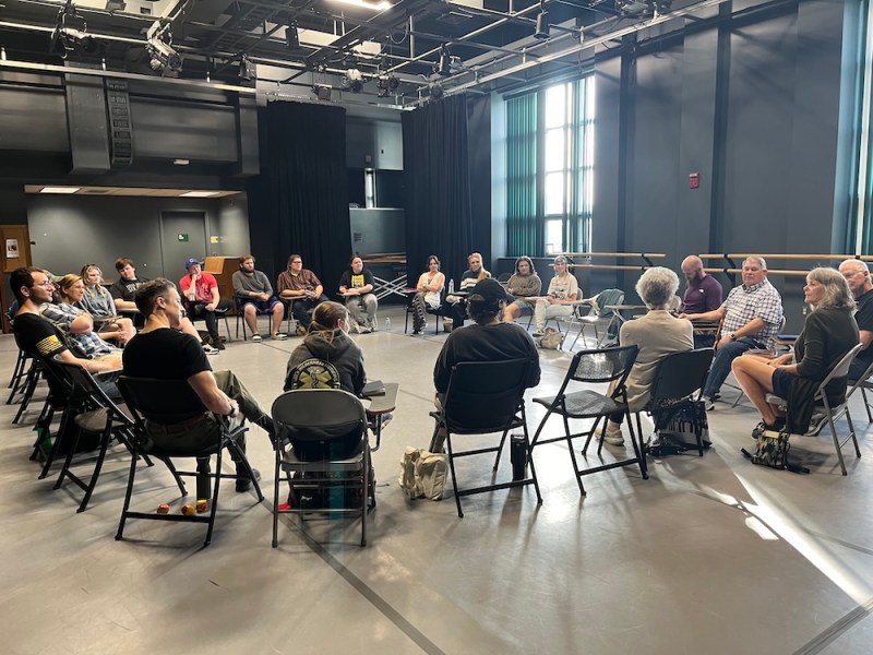 A group of people seated in a large circle during a discussion in a theater rehearsal space.