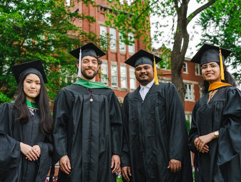 Four graduates stand wearing graduation caps and gowns in front of an old brick building