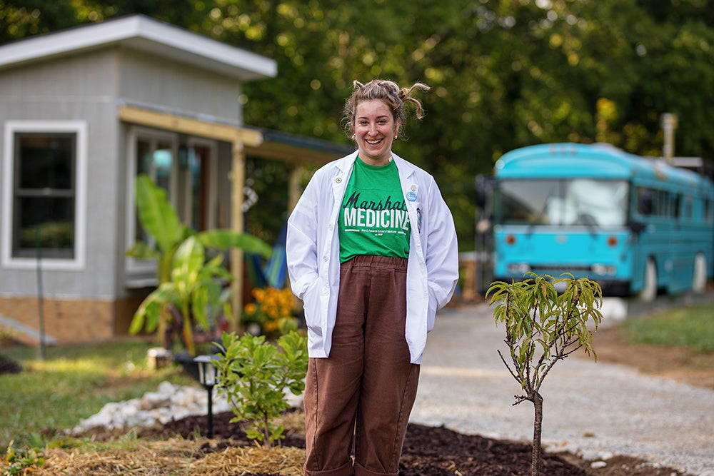 Carina Raya wears a medical school t-shirt and white coat while standing in front of her tiny home
